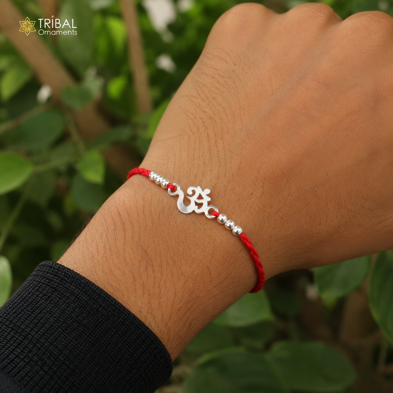 Sterling silver OM design bracelet with red thread rakhi on wrist, showcasing intricate jewelry details against leafy background.
