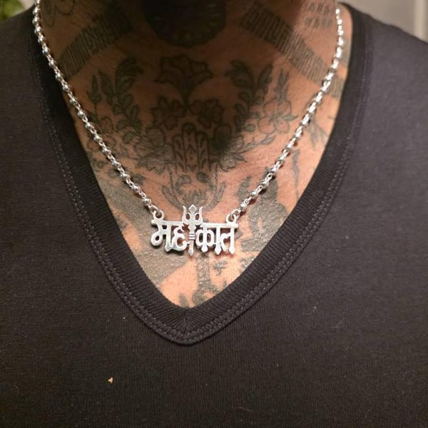 A person wearing a silver necklace with mahakal name on a black shirt with tattoos.